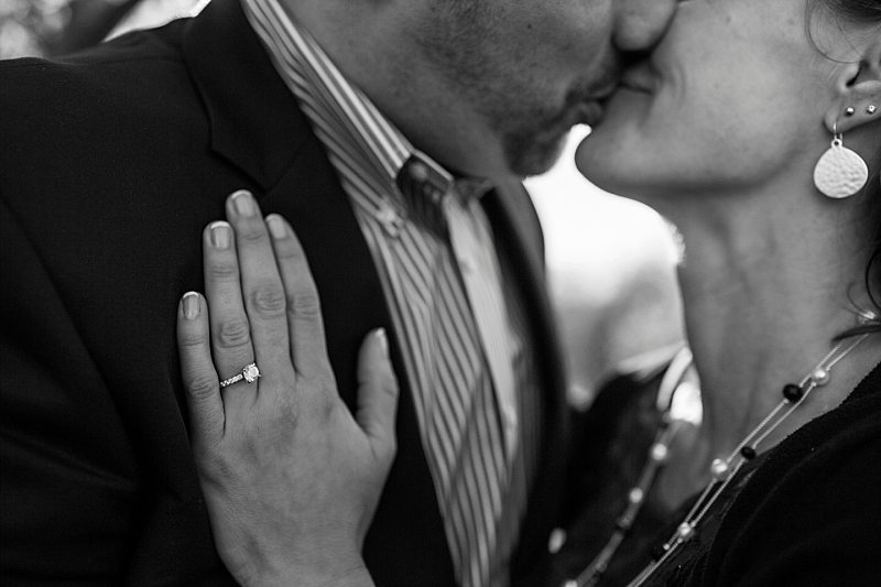 A black and white close-up of an engagement ring as the bride and groom kiss in the background.
