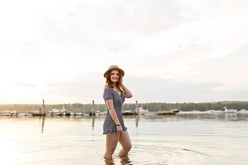 A woman stands in the ocean wearing a romper and hat at sunset.