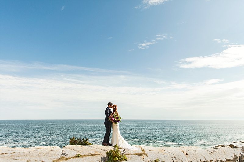 A portrait of a bride and groom kissing on the cliffs at Pemaquid Point Light on a sunny day.