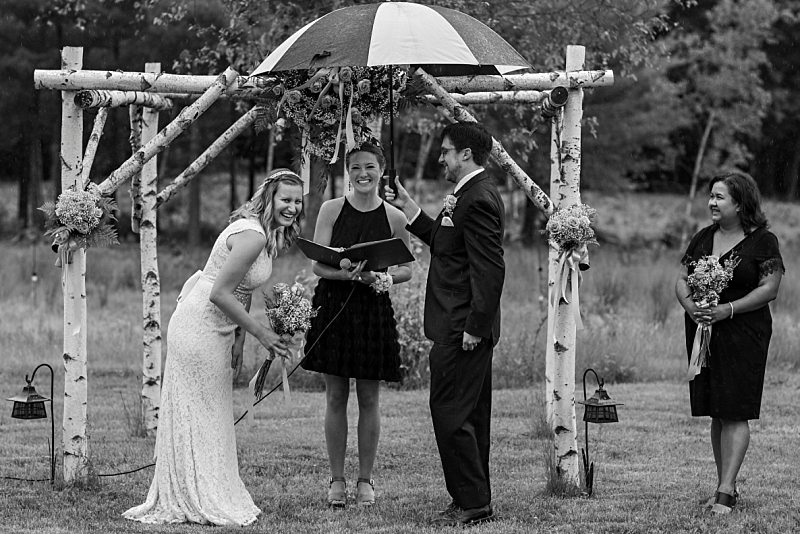 A bride and groom laugh as it rains during their ceremony. The groom holds an umbrella over them.