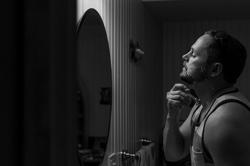 A dramatic black and white photo of a groom shaving his beard while looking into a mirror.