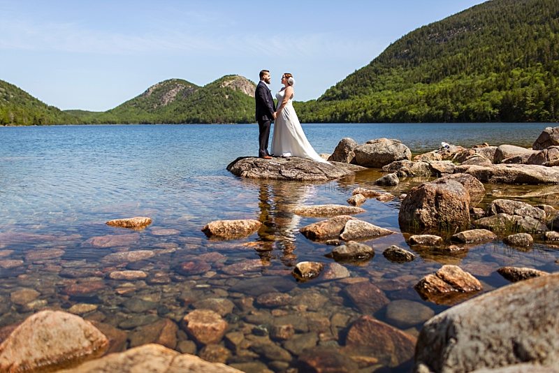 A portrait of a bride and groom on a sunny day at Jordan Pond in Acadia National Park. They stand on rocks jutting out into the water, holding hands.