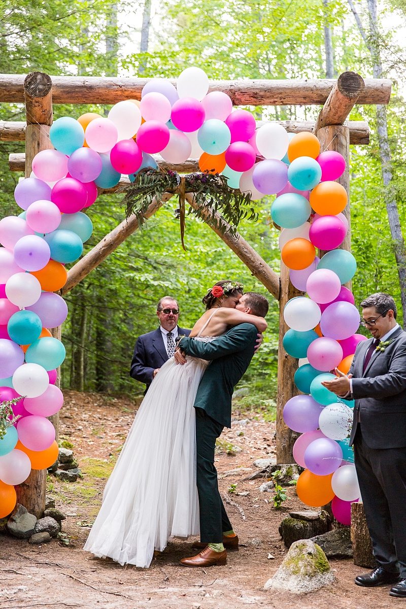 A groom lifts the bride during their first kiss under a balloon-covered arbor.