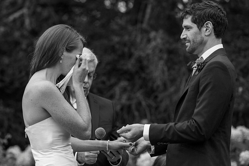 A black and white photo of a bride wiping her tears with the handkerchief the groom has just handed her during the ceremony.