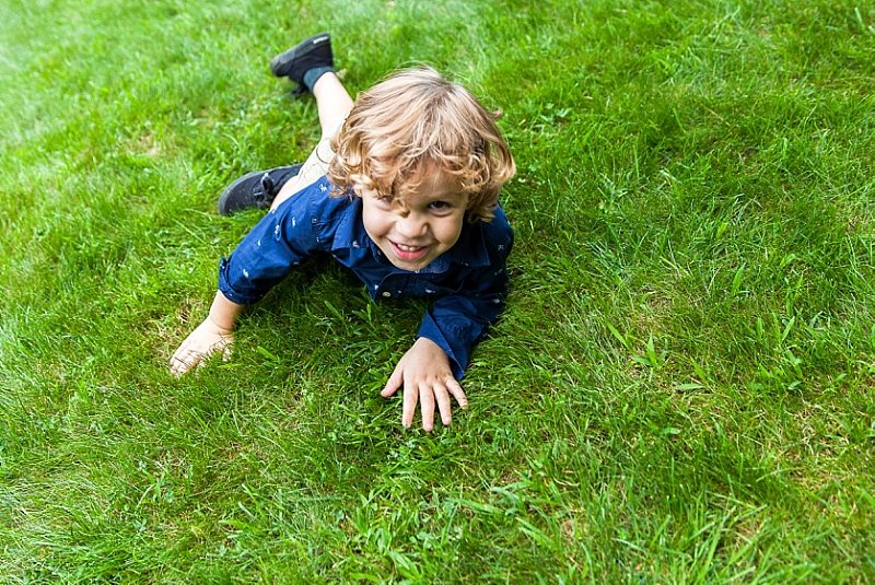 A little boy at a wedding crawls in the grass and smiles at the camera.
