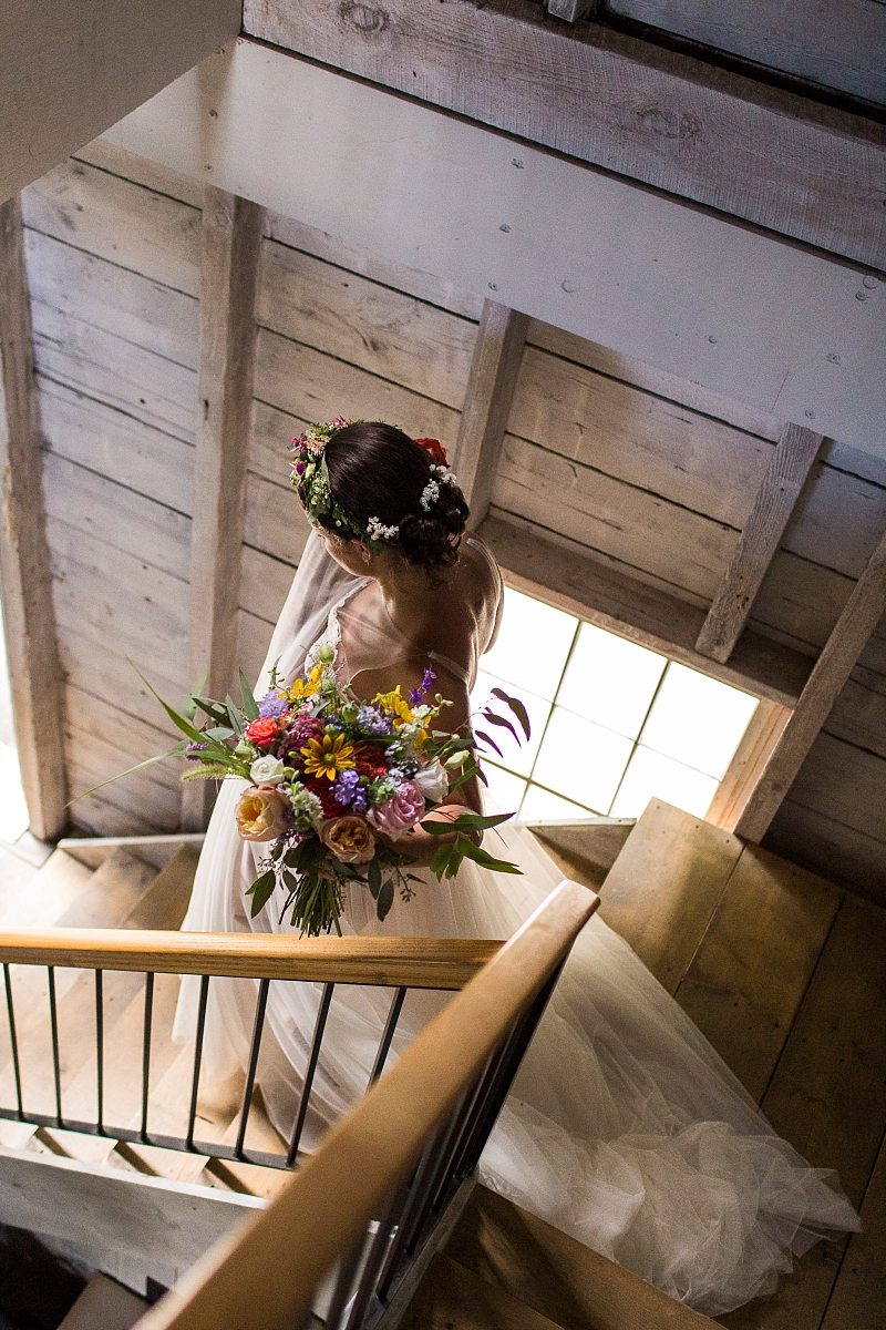 A bride descends the stairs in a beautiful, white-washed barn carrying her bouquet with sunlight falling on her.