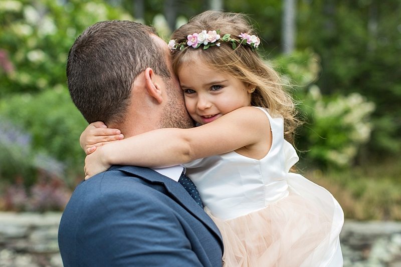 The groom carries the flower girl as she hugs him tightly around the neck.