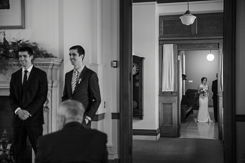 A black and white image of the bride waiting to enter the ceremony down the hall. The groom can be seen waiting and is slightly out of focus.