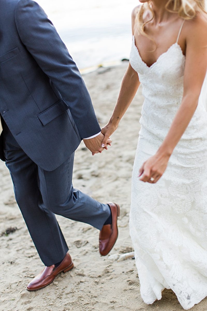 A close up of a bride and groom's hands. They hold hands as they walk together on the beach.