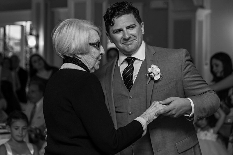 An emotional black and white photo of a groom crying as he dances with his grandmother at his wedding reception.