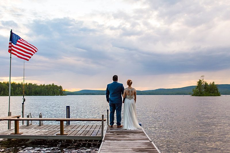 A bride and groom's backs as they face sunset on a dock with an American flag waving in the wind nearby.