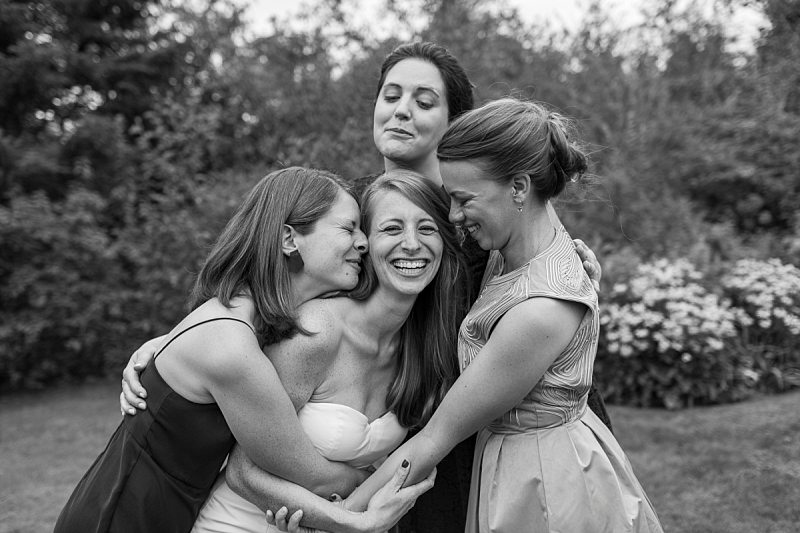 Bridesmaids tightly hug the bride and she laughs.