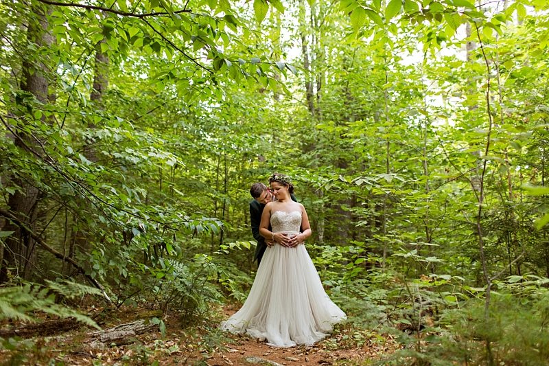A portrait of a bride and groom standing in the Maine wilderness with their arms around each other.