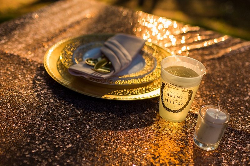Sunlight shines on the tablecloth, place setting and through a cup creating a golden glow. The cup states the name of the couple and their wedding date.