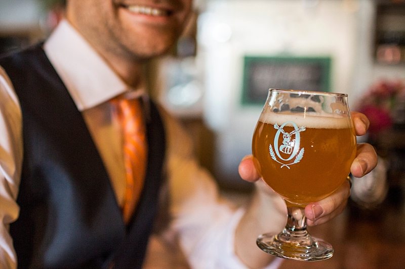 You can see a groom smiling in the background of this close-up of the beer he is holding.