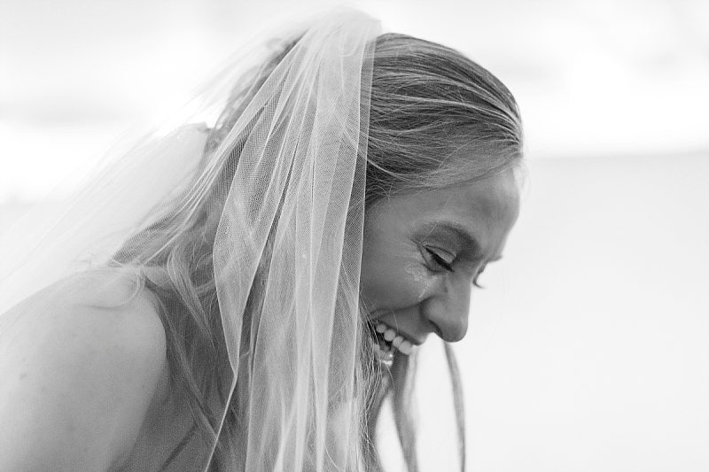 A black and white of a bride laughing with tear-stained cheeks during her wedding ceremony.