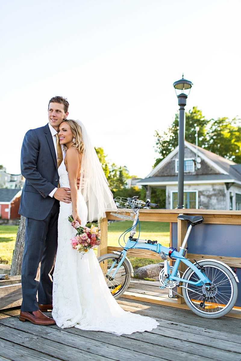 A bride and groom stand on a dock, looking out at the ocean, with a blue bicycle standing nearby.