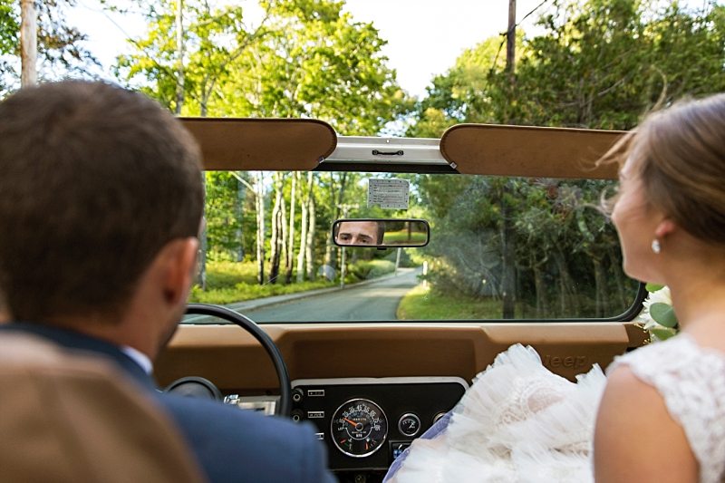 The bride and groom ride together as they head out for wedding portraits. The focus is on the groom's eyes in the rear-view mirror.