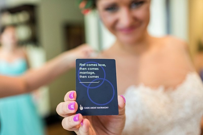 A bride holds a card out at the camera that says, "first comes love, then comes marriage, then comes (blank)."
