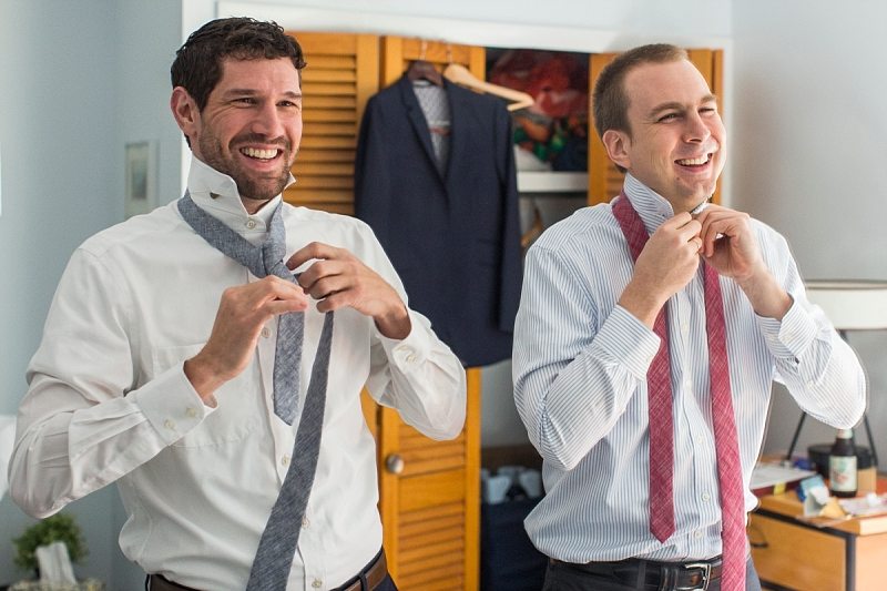 A groom and his best man laugh as they struggle to tie their ties.