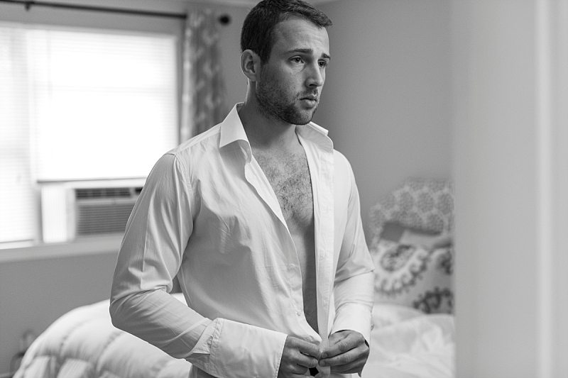 A groom buttons up his shirt as he looks in the mirror.