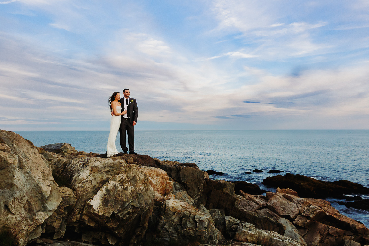 Bride and groom stand on cliffs at Otter Point in Acadia National Park at sunset.