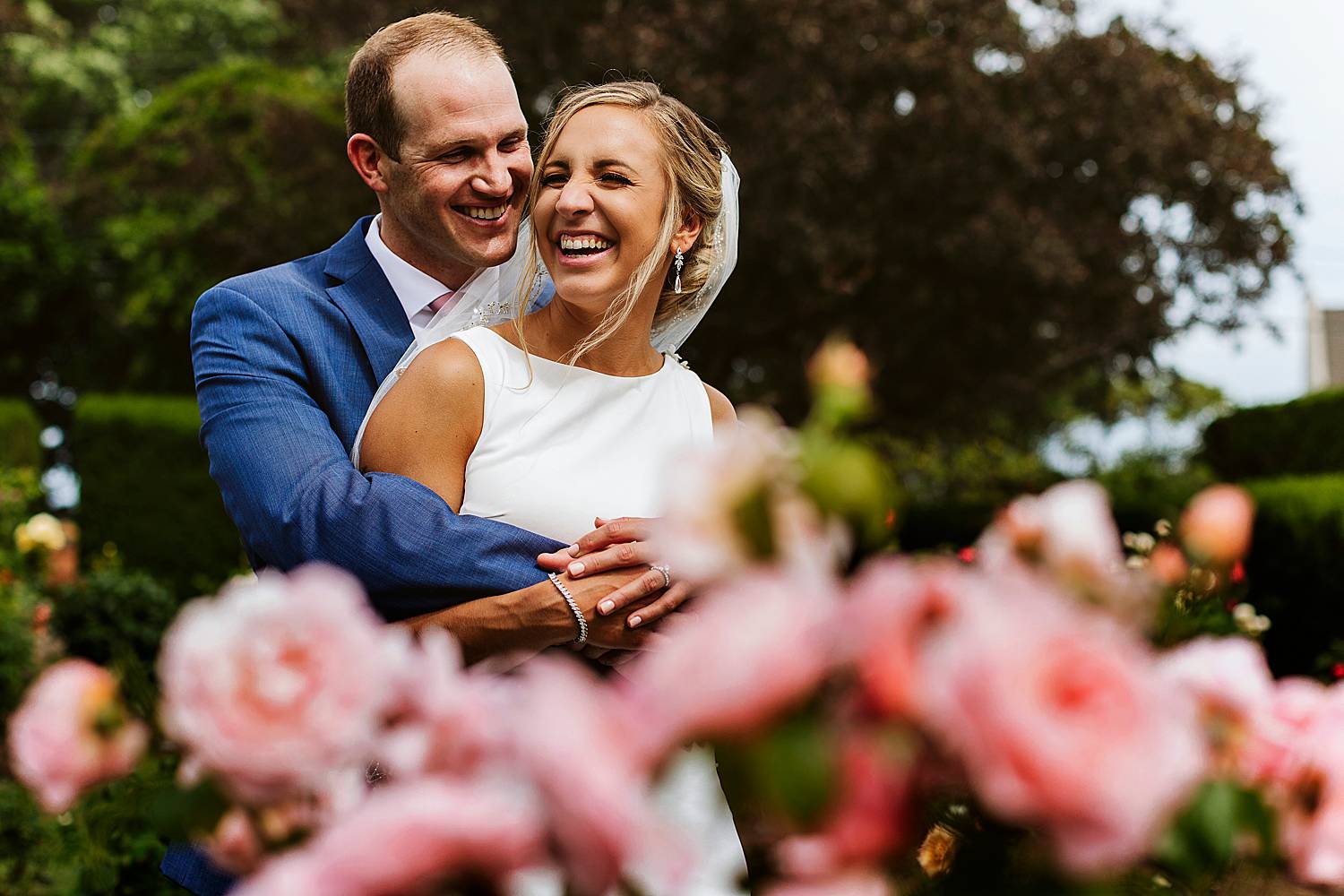 bride and groom laughing together behind roses at North Hampton, NH wedding at fuller gardens